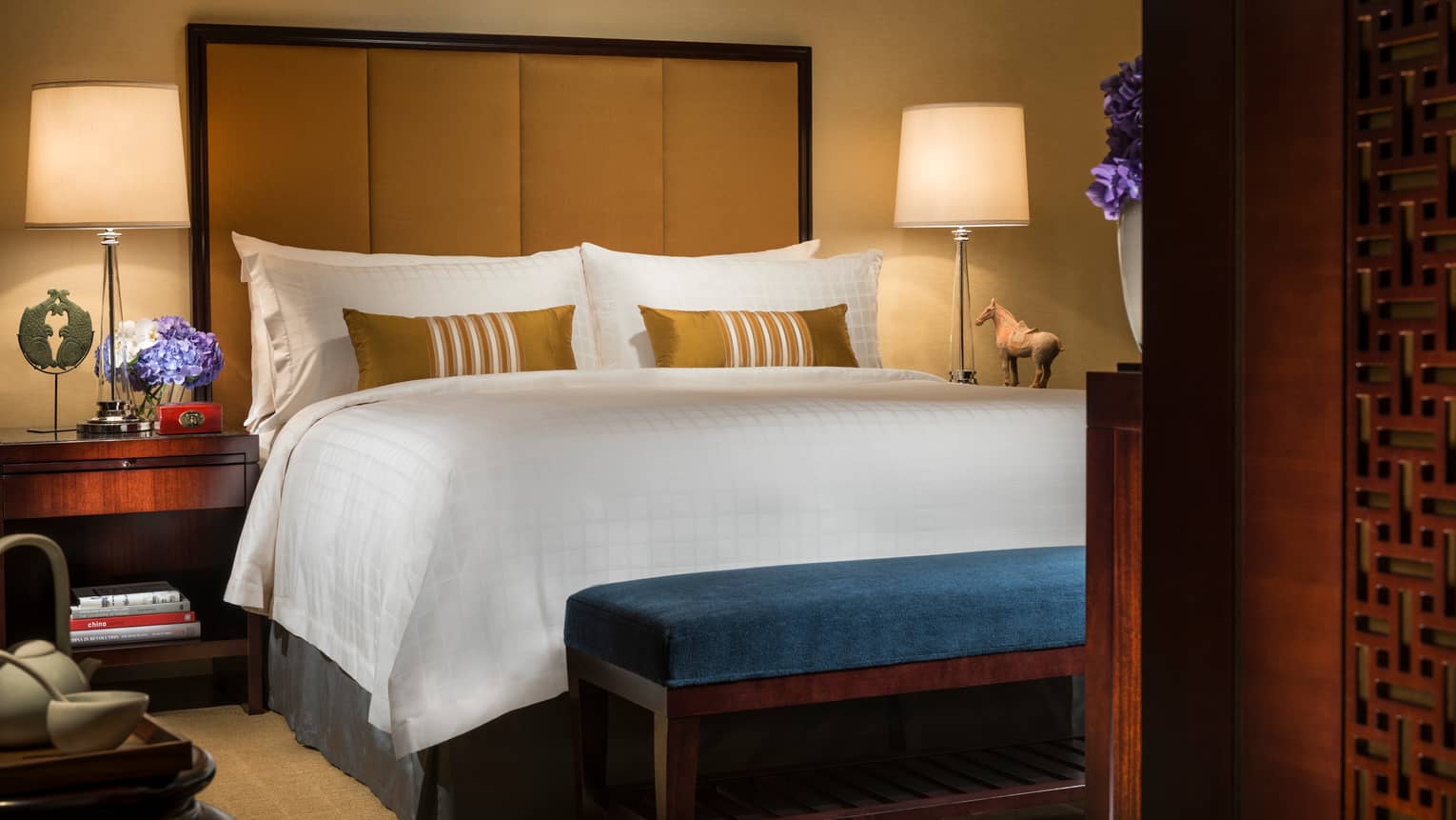 Deluxe Room bed with tall padded headboard, small blue bench at foot, sculptures and art on nightstand