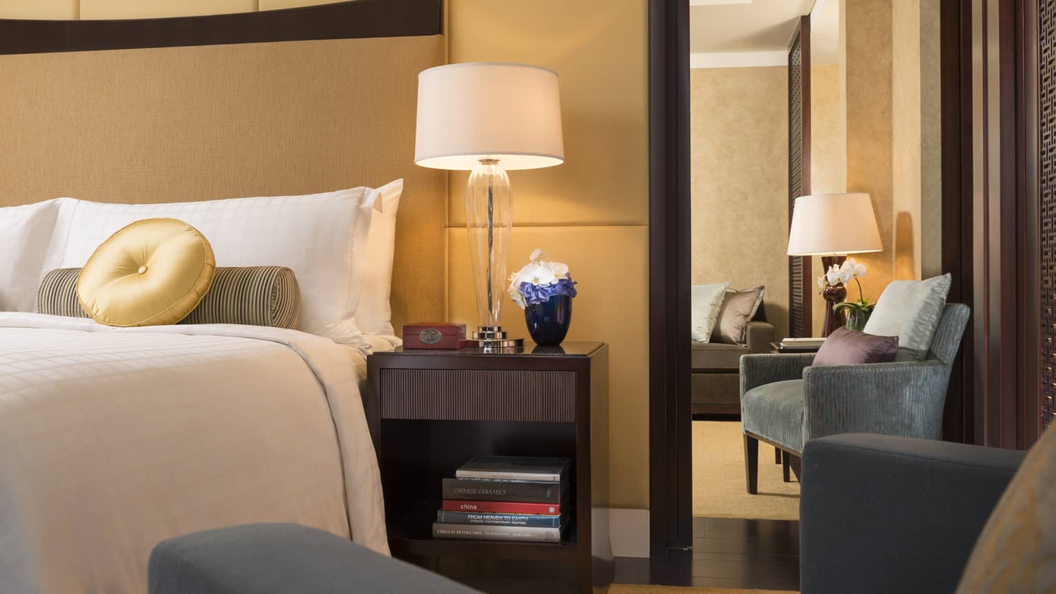 Ambassador Suite corner of bed with round gold pillow, armchair, nightstand with lamp, flowers
