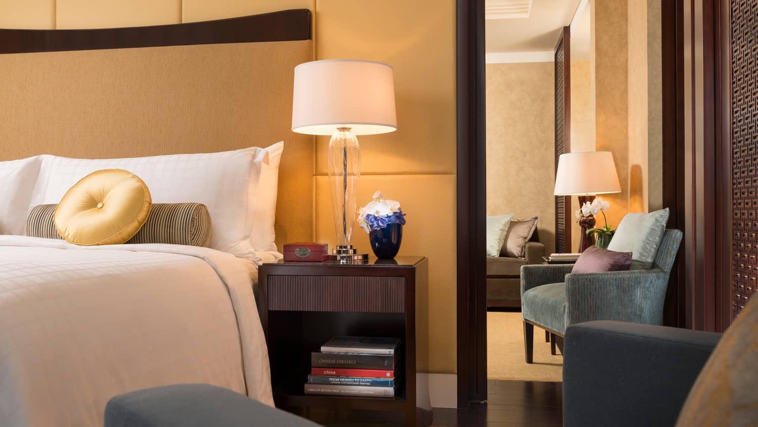 Ambassador Suite corner of bed with round gold pillow, armchair, nightstand with lamp, flowers