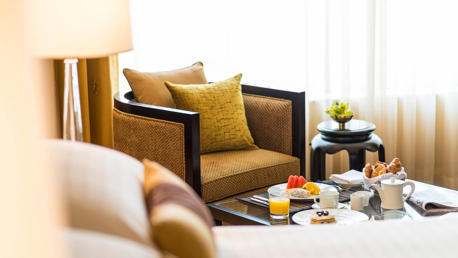 Gold armchair, pillow by in-room dining table with fresh fruit, pastries, coffee, juice