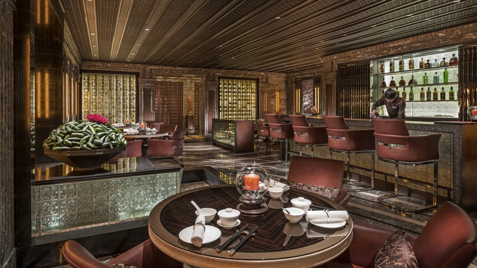 Bartender at bar lined with brown stools behind small round lounge tables, chairs in dimly-lit room with dark wood details