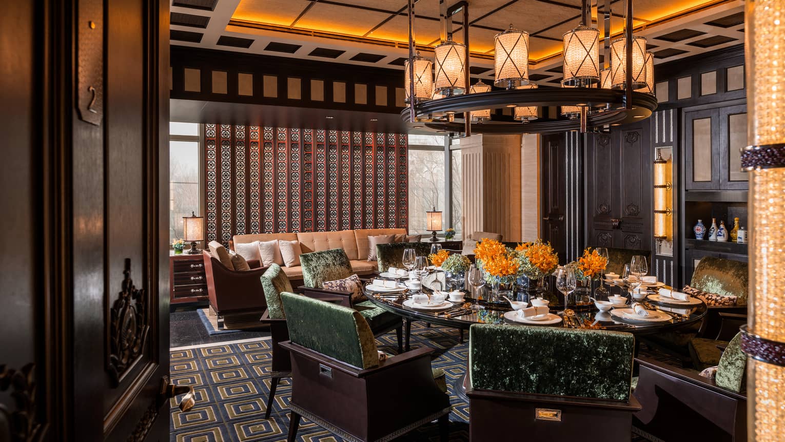 Large round dining table with green crushed-velvet chairs, orange flowers, wine glasses, under large chandelier