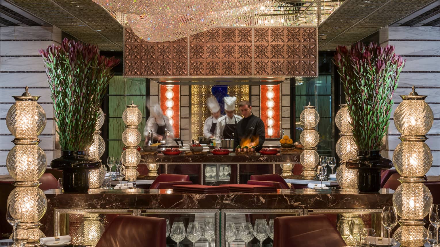 Chefs in open kitchen, flames from pot in front of black marble tables with tall crystal lamps