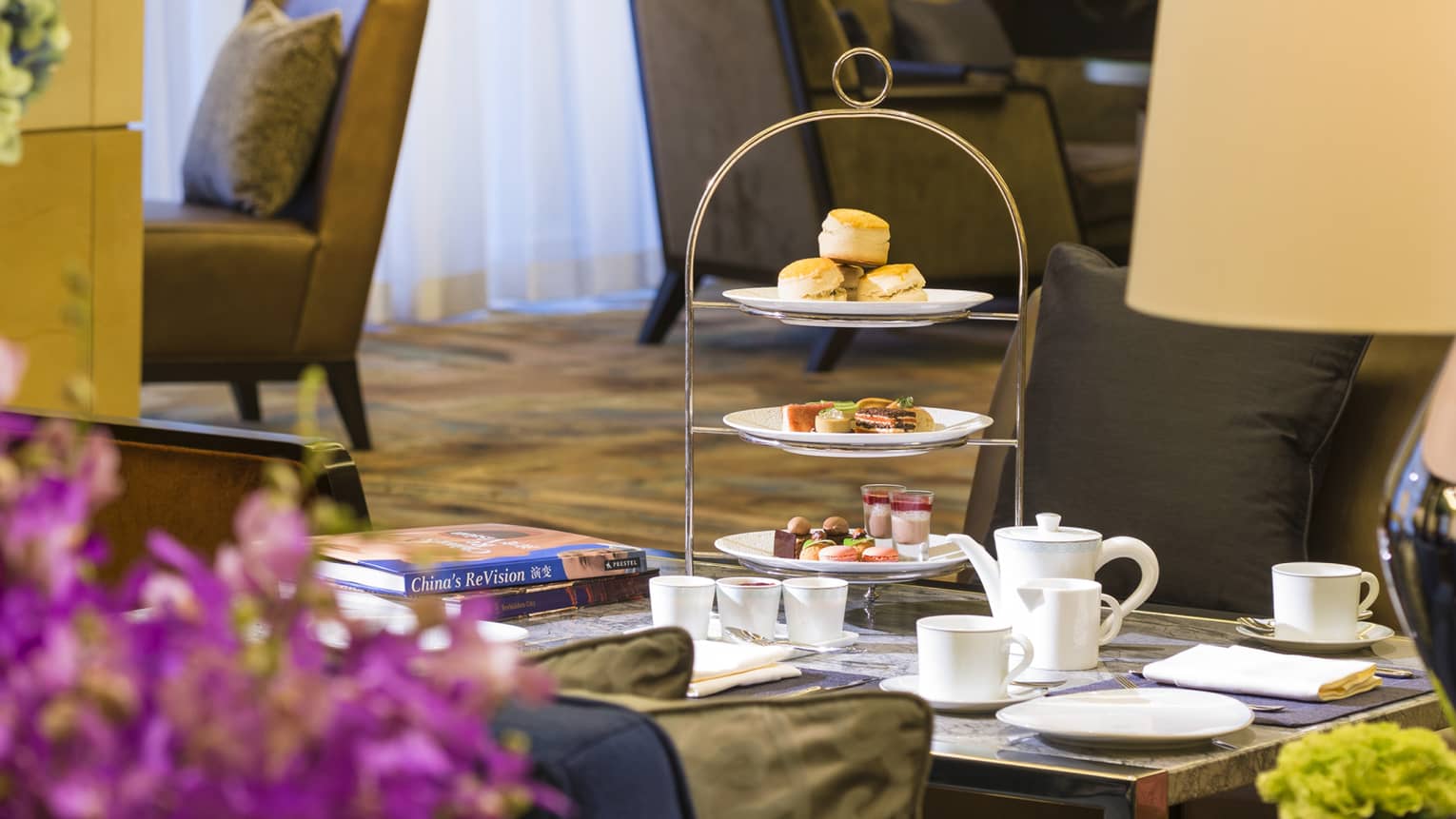 Tray with afternoon tea desserts, teapot service on coffee table past fresh purple flowers