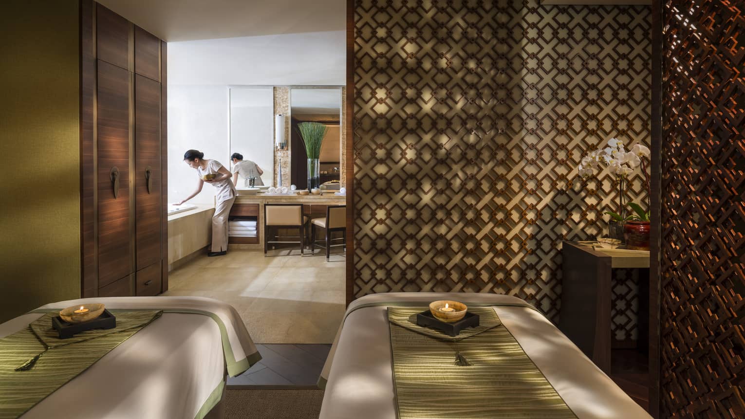 Couples massage beds side-by-side in spa room with candles, wood detail wall, staff preparing bath