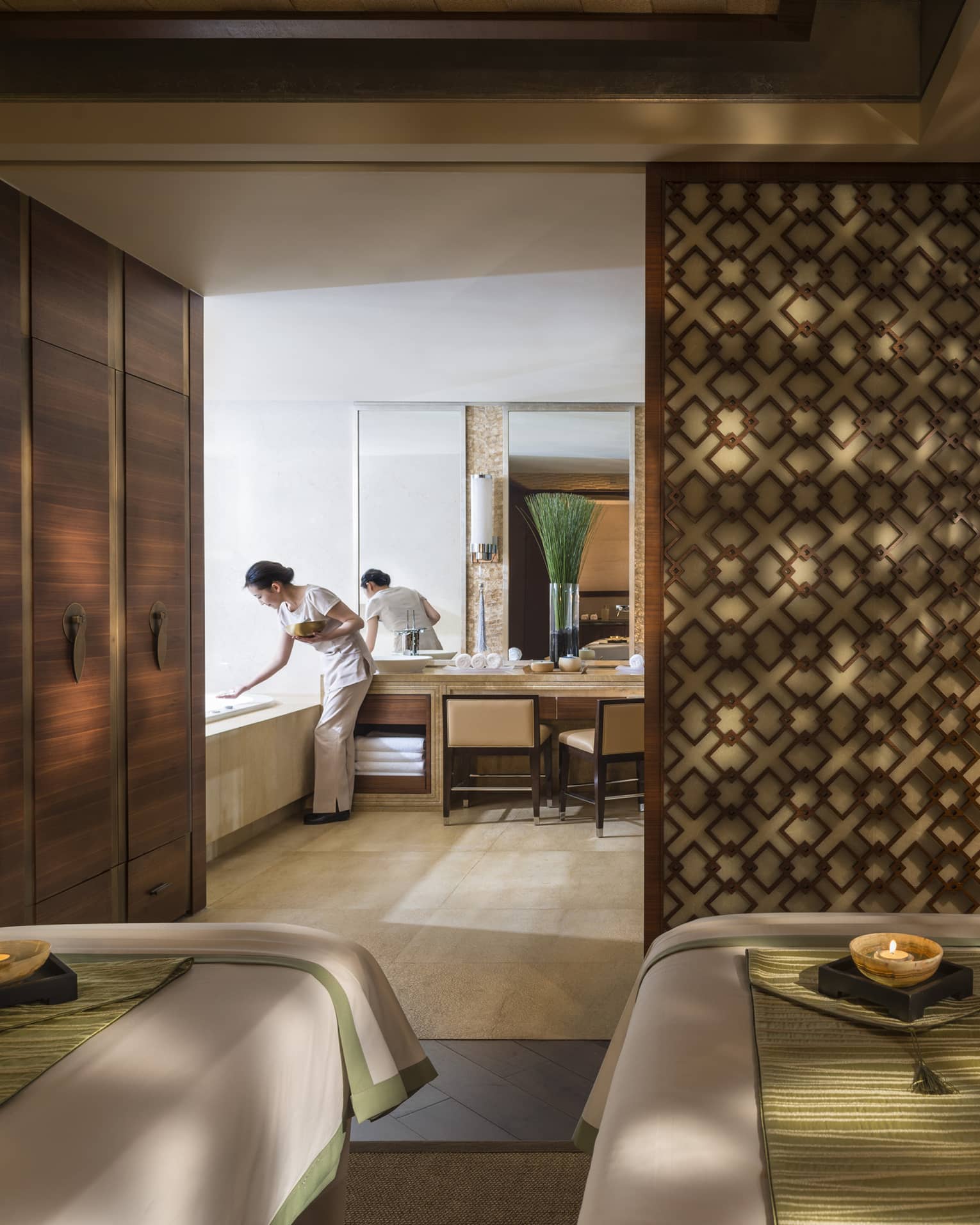 Couples massage beds side-by-side in spa room with candles, wood detail wall, staff preparing bath